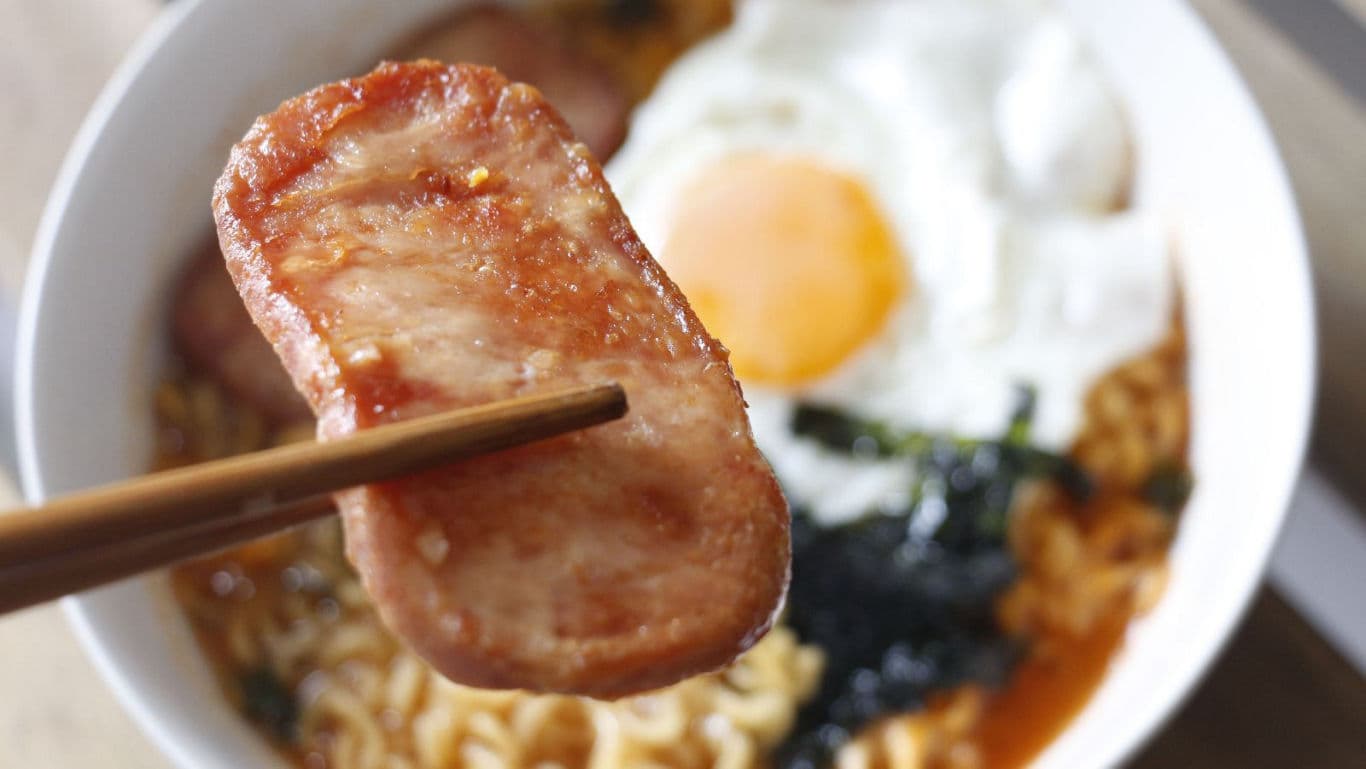 Chopsticks holding spam over ramen bowl