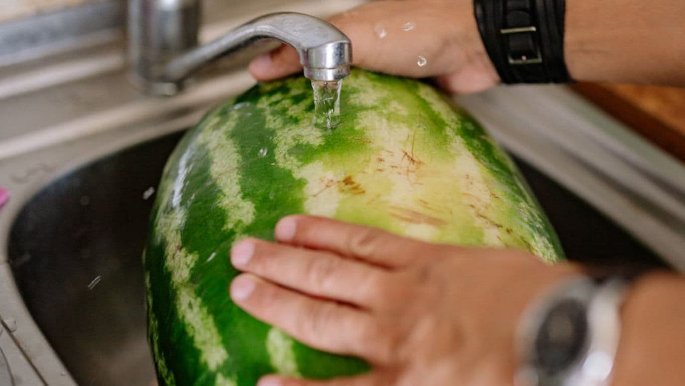 Person washing large watermelon under kitchen faucet