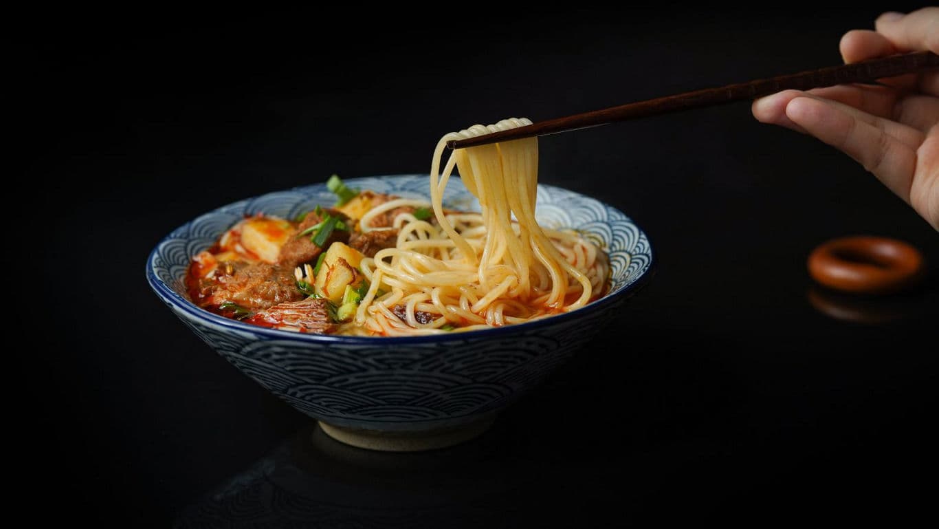 Hand lifting noodles from steaming soup bowl